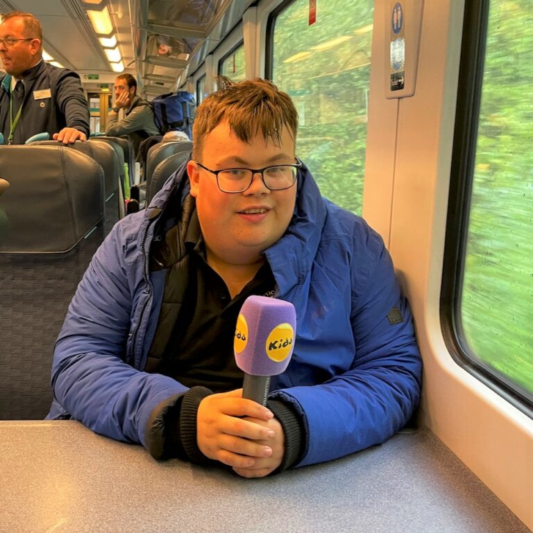 Charlie sits on a train looking relaxed and holding a microphone.